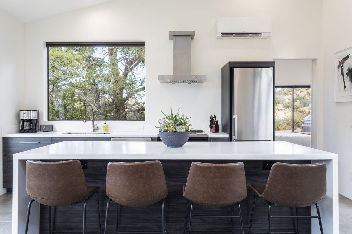 Una cocina moderna con una isla grande, taburetes de bar, un ventanal con vistas a árboles y una campana extractora de acero inoxidable.