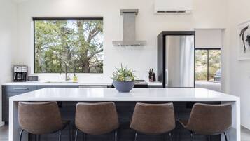 Una cocina moderna con una isla grande, taburetes de bar, un ventanal con vistas a árboles y una campana extractora de acero inoxidable.