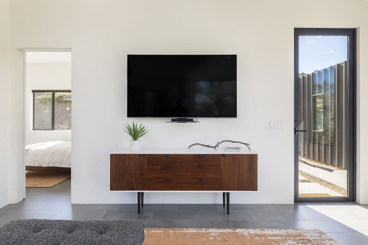 En moderne stue med et fladskærms TV monteret på en hvid væg, et træsofa bord med skuffer, en lille potteplante og en stor skydedør i glas, der fører til et udendørs område.
