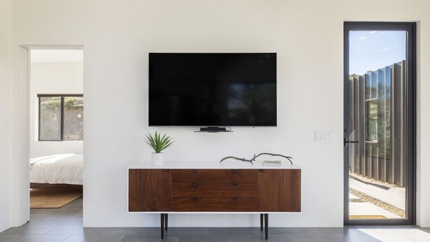 Un salon moderne avec un téléviseur à écran plat fixé sur un mur blanc, une console en bois dotée de tiroirs, une petite plante en pot et une grande porte-fenêtre coulissante donnant sur un espace extérieur.
