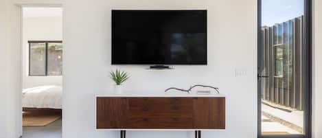 Una sala moderna con un televisor de pantalla plana montado en una pared blanca, una consola de madera con cajones, una pequeña planta en maceta y un amplio ventanal corredero que da a un área exterior.
