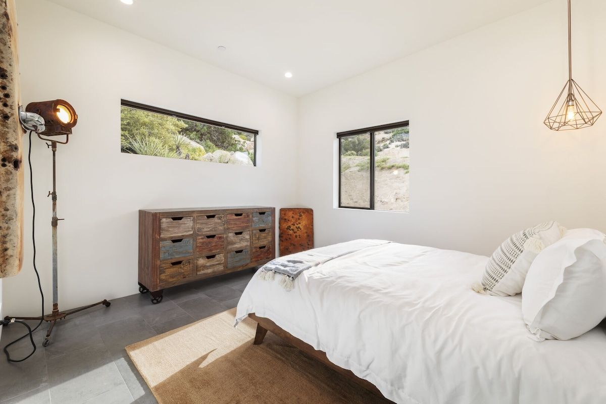 Une chambre avec un grand lit, une commode en bois, une lampe sur pied et deux fenêtres donnant sur un paysage verdoyant.
