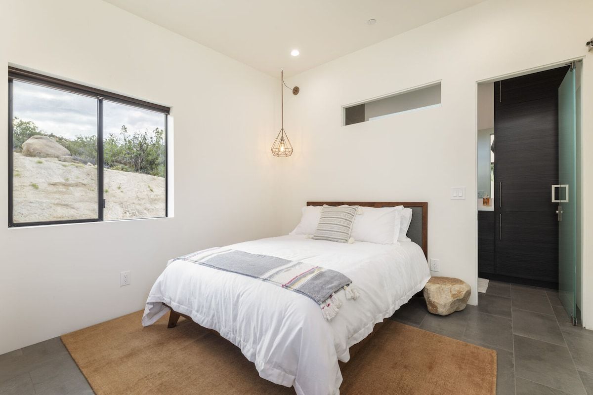 Un dormitorio con cama, ventana con vista a paisaje desértico, lámpara colgante y puerta que da a otra habitación.