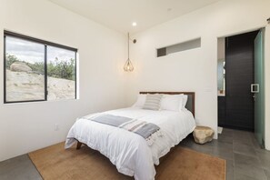 A bedroom with a bed, a window with a view of a desert landscape, a hanging light fixture, and a door leading to another room.