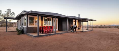 Un edificio moderno de una sola planta con un patio cubierto, una mesa de comedor con sillas y una parrilla de barbacoa.