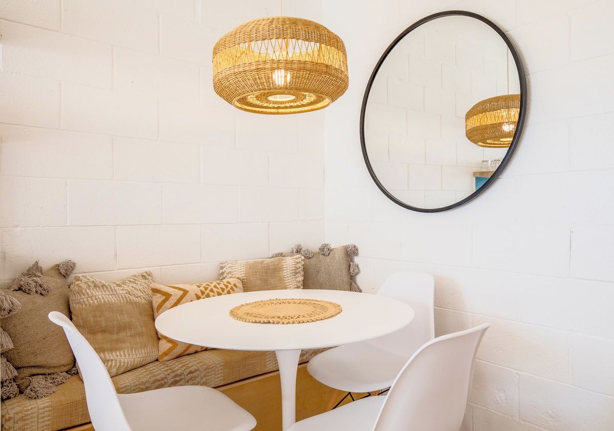 A room with a white brick wall, a round table with chairs, a woven pendant light, and a round mirror.