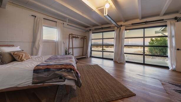 A bedroom with a large bed, a view through a sliding glass door, and a ceiling light fixture. - That Desert House - Hot Tub, Privacy & Desert Views! (Twentynine Palms)