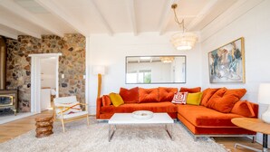 A living room with a stone wall, a white sofa with orange cushions, a white coffee table, and a wooden side table. - That Desert House - Hot Tub, Privacy & Desert Views! (Twentynine Palms)