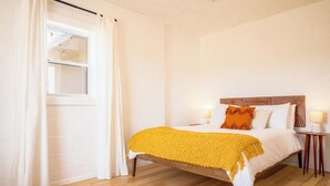 Une chambre à coucher avec un lit, une fenêtre avec des rideaux, une table de chevet avec une lampe et un plancher en bois.