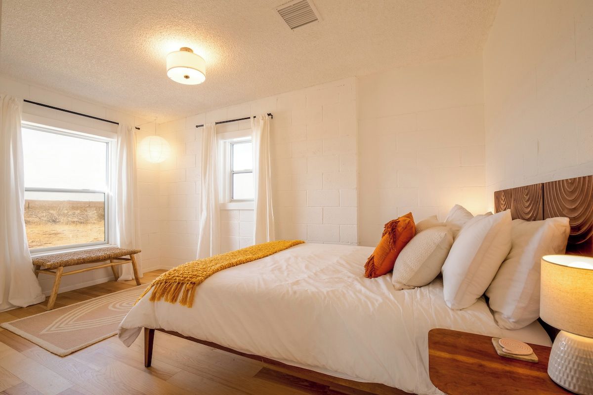 Une chambre à coucher avec un grand lit, un banc, une table de chevet et une fenêtre avec des rideaux.