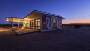 A modern house with a large glass door, a covered patio, and a desert landscape in the background. - That Desert House - Hot Tub, Privacy & Desert Views! (Twentynine Palms)