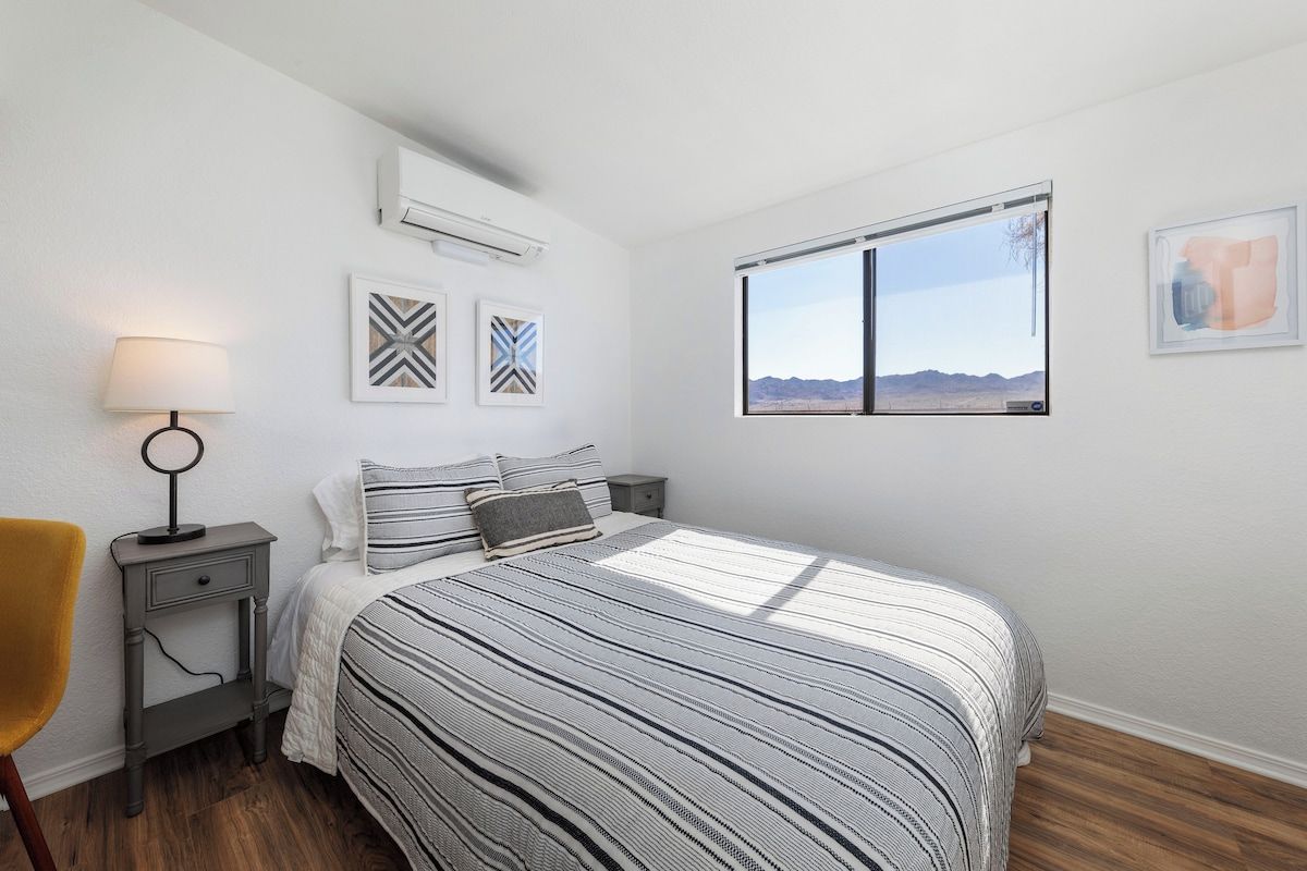 A bedroom with a bed, a nightstand, a lamp, a window with a view, and wall art.