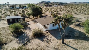 A remote desert house with a small attached garage, surrounded by sparse vegetation and distant hills. - Mojave Mesa - Secluded Desert Home Near Joshua Tree (Yucca Valley)