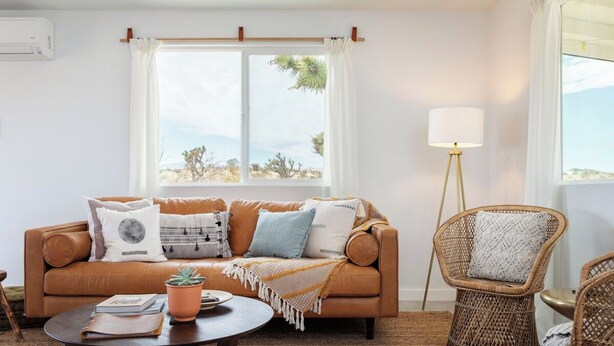 A living room with a brown sofa, a round coffee table, a potted plant, and a wicker chair. - Mojave Mesa - Secluded Desert Home Near Joshua Tree (Yucca Valley)