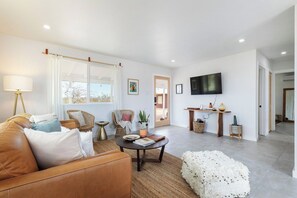 Un salon avec un canapé en cuir, une table basse ronde et un pouf blanc. Un téléviseur est fixé au mur et il y a une fenêtre avec des rideaux.