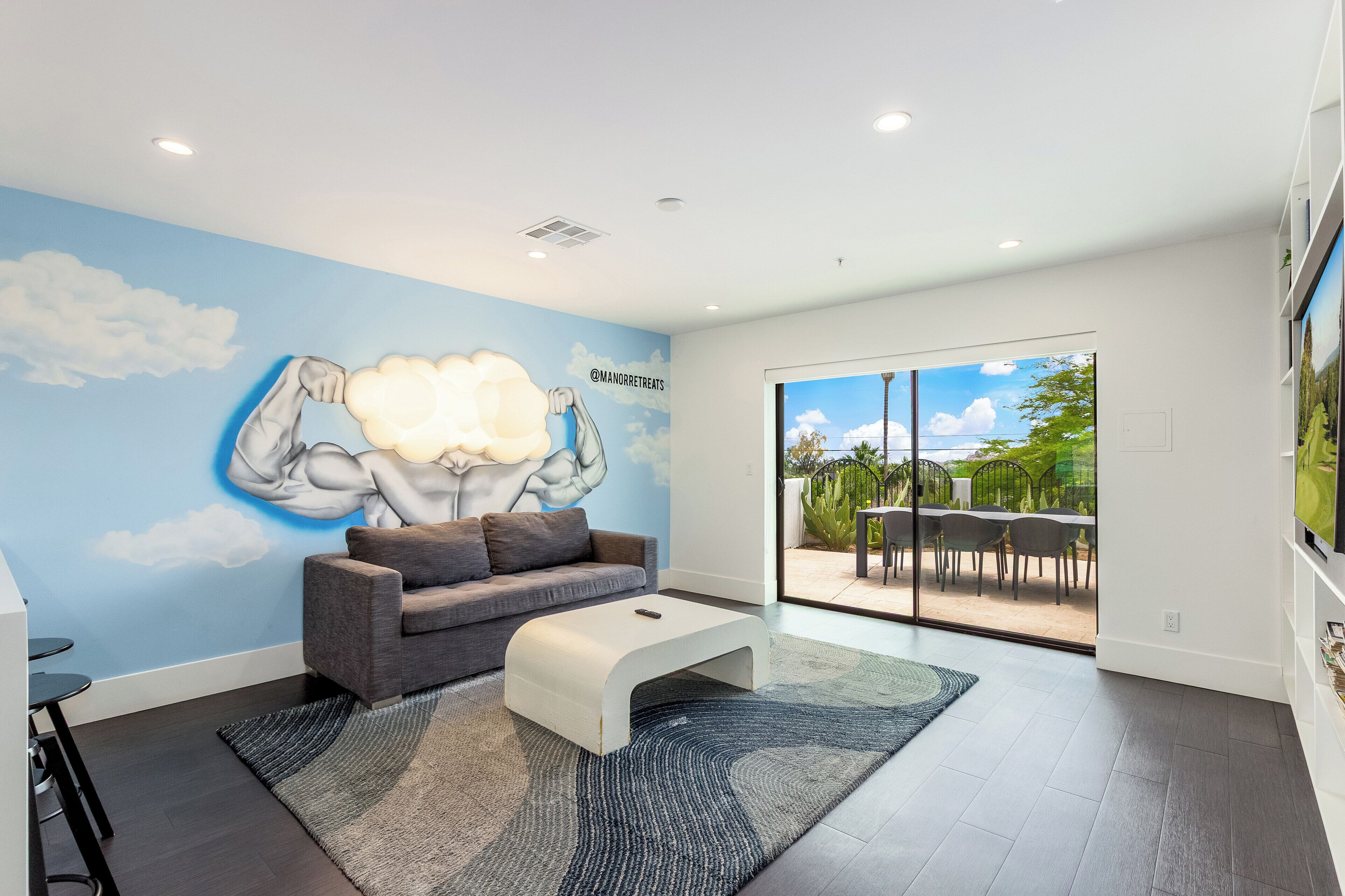 A modern living room with a mural, a gray sofa, a white coffee table, and a large sliding glass door leading to an outdoor patio.
