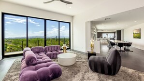 Un vaste espace de vie avec une grande fenêtre, un ventilateur de plafond, une table basse ronde et, au fond, une cuisine moderne.