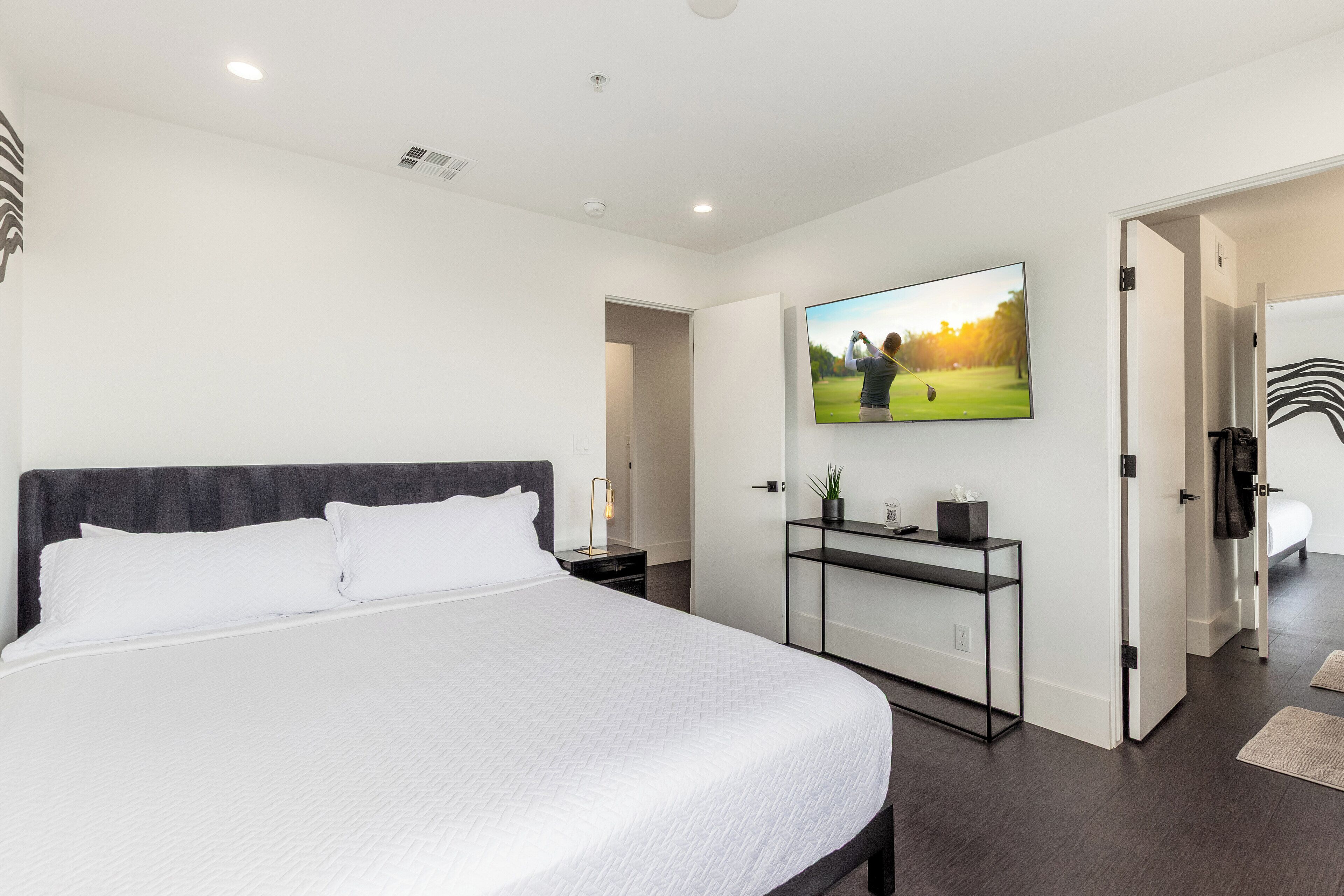 Une chambre moderne avec un grand lit, un téléviseur fixé au mur et une porte donnant sur une autre pièce.