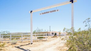 A wooden gate with a sign reading 'FLAMINGO RANCH' in a desert landscape with sparse vegetation and a clear sky. - Flamingo Ranch - Dreamy Desert Design (Yucca Valley)
