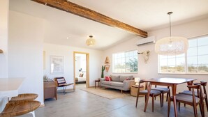 A spacious living area with a dining table, wooden chairs, a sofa, and a large window providing natural light. - Flamingo Ranch - Dreamy Desert Design (Yucca Valley)