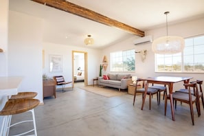 A spacious living area with a dining table, wooden chairs, a sofa, and a large window providing natural light.