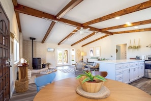 A spacious living area with a wooden ceiling, a dining table with a potted plant, and a kitchen with white cabinets.