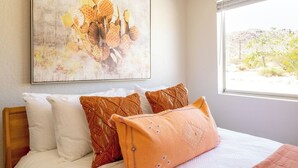 A bed with white and orange bedding, a large window with a view of a desert landscape, and a wall-mounted abstract painting.