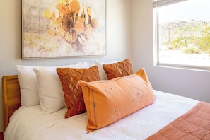 A bed with white and orange bedding, a large window with a view of a desert landscape, and a wall-mounted abstract painting.