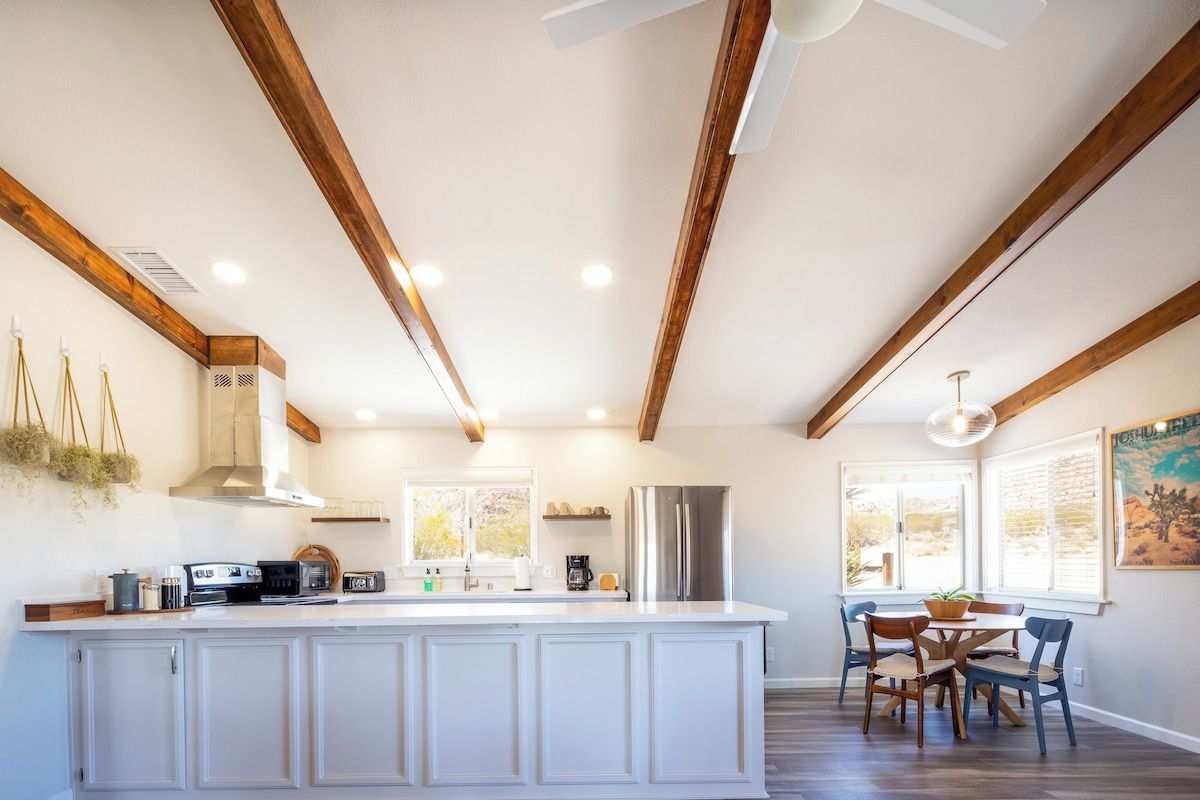 Una cocina con isla central, vigas de madera, iluminación empotrada y un comedor con sillas y una mesa.