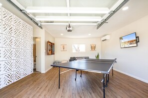 A room with a ping pong table, wooden flooring, a white partition with a geometric pattern, a ceiling with skylights, and a mounted TV.