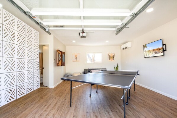 A room with a ping pong table, wooden flooring, a white partition with a geometric pattern, a ceiling with skylights, and a mounted TV.