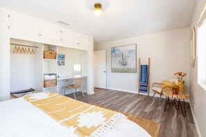 Un dormitorio con cama, escritorio y silla. Se observa un armario con ropa colgada y una estantería con cestas. Un cuadro adorna la pared, y una lámpara está sobre el escritorio.