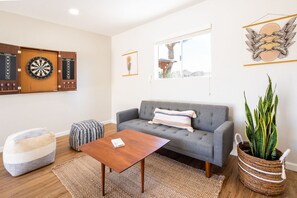 A living room with a grey sofa, a wooden coffee table, a woven pouf, a dartboard, and a potted plant.
