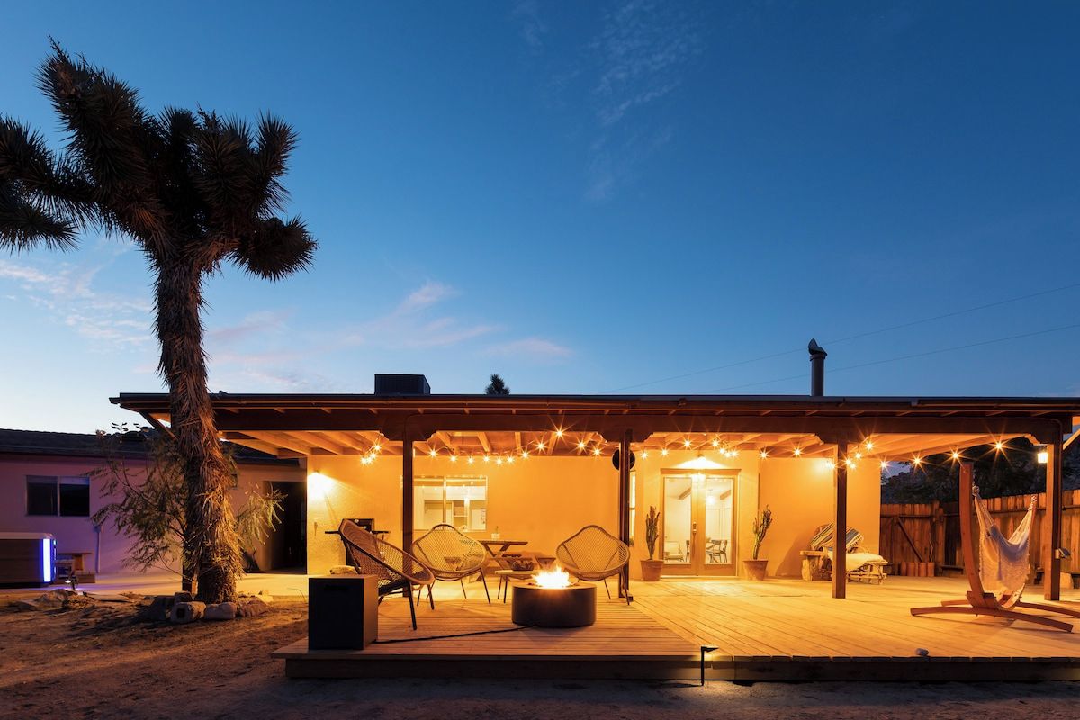 Un patio techado al aire libre con mobiliario y una chimenea, rodeado por una terraza de madera y una palmera.