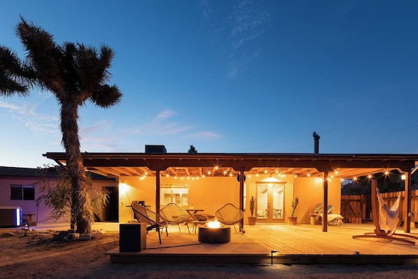 A covered outdoor patio with seating and a fire pit, surrounded by a wooden deck and a palm tree.