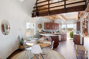 A modern kitchen with a central island, wooden ceiling beams, and a dining area with a round table and chairs. - Abracadabra Dome in the Desert-Unique Experience! (Landers)