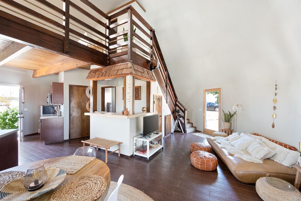 Un vaste espace de vie comprenant un escalier en bois, un coin cuisine et un espace détente avec un canapé et des poufs.
