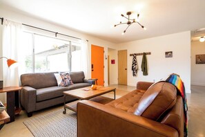 Una sala de estar moderna con un sofá, una mesa de centro y un sillón de cuero. Se ve una ventana con cortinas, una puerta y un perchero de pared.