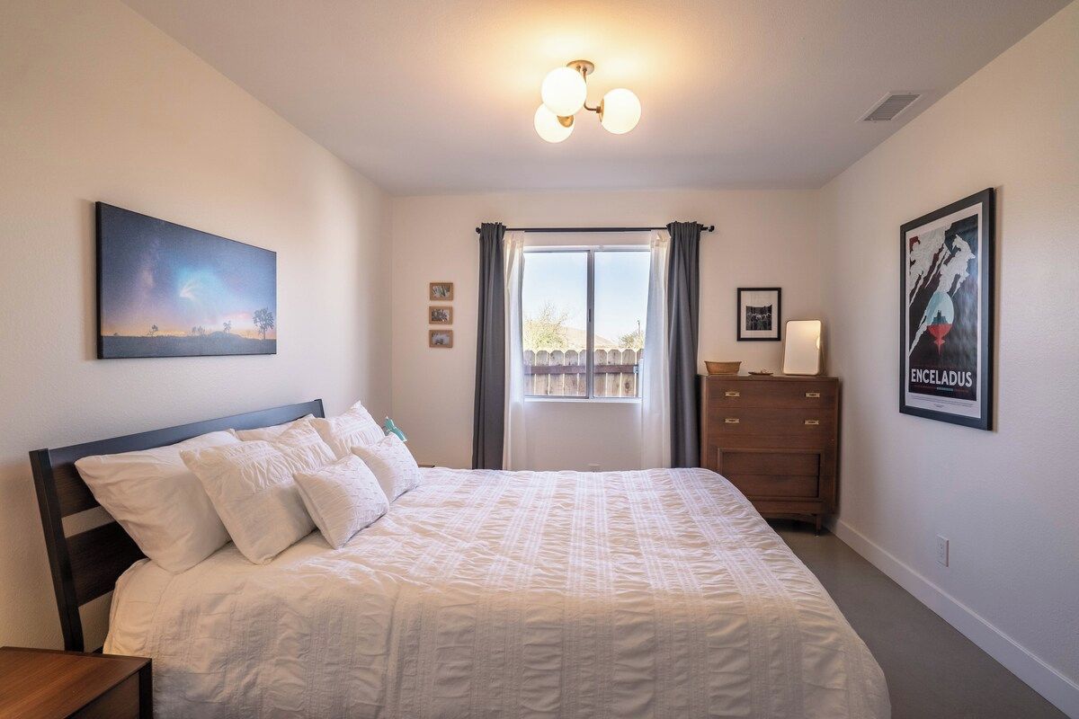 Ein Schlafzimmer mit Bett, Kommode, einem Fenster mit Vorhängen und Wandbildern.
