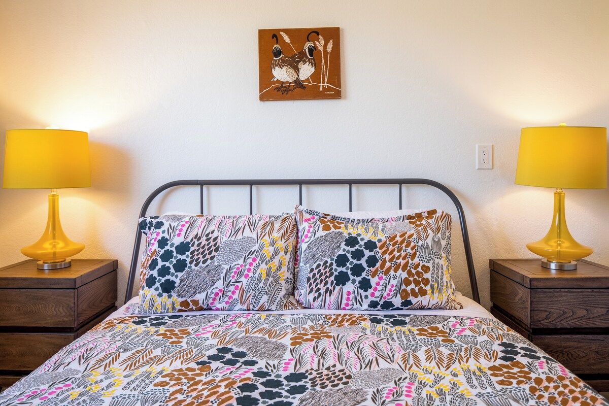 A bed with patterned bedding, two bedside lamps, a wall-mounted artwork, and a power outlet.