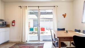 A dining area with a wooden table and chairs, a fire extinguisher on the wall, and a view through a glass door.