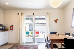 A dining area with a wooden table and chairs, a fire extinguisher on the wall, and a view through a glass door.