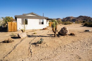 A single-story house with a white exterior, a wooden fence, and a desert landscape with cacti and rocks.