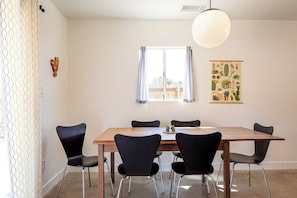 Uma área de refeições com mesa de madeira e cadeiras pretas, uma janela com cortinas e ilustração de cacto na parede.