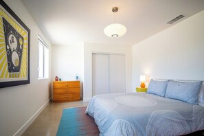 Un dormitorio con cama, cómoda, mesita de noche con lámpara, puerta y un cuadro en la pared.