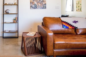 Um sofá de couro marrom com uma almofada vibrante de padrão geométrico, uma mesinha de madeira com um livro e uma estante de metal preto com diversos objetos.
