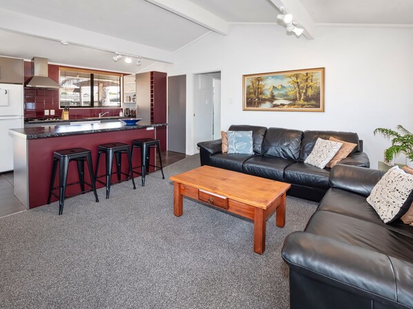 A modern living room with a black leather sofa, a wooden coffee table, and a kitchen area with a red island and black stools.