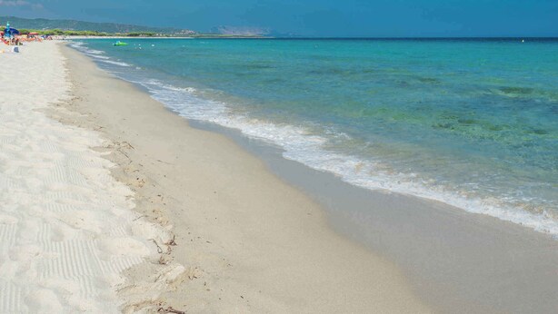 Una playa con aguas turquesas cristalinas, orilla de arena y colinas al fondo.