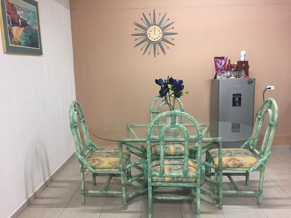 A dining area with a glass table surrounded by four green chairs with patterned cushions. A decorative wall clock and a water dispenser are visible in the background.