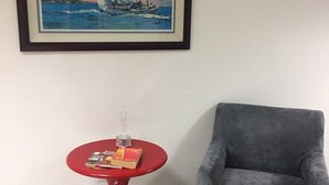 A room with a framed painting of sailboats, a red round table with a glass, and a grey armchair.